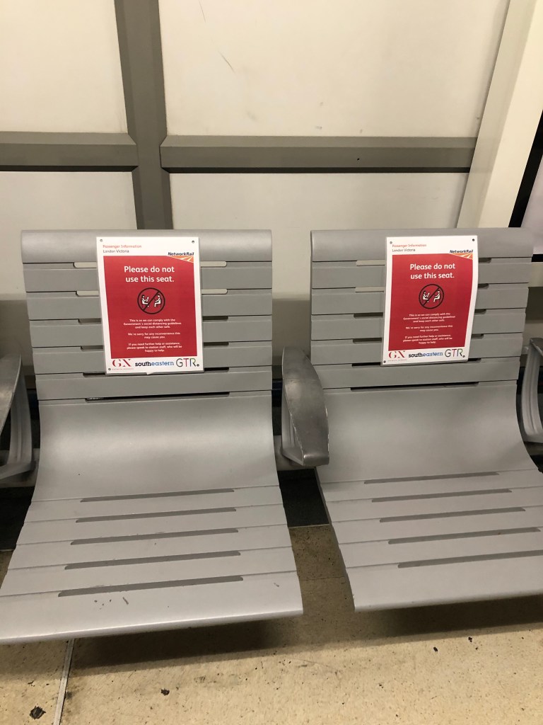 Two grey train station benches both with red signs warning people not to sit on them due to social distancing 